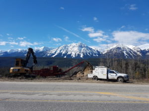 A truck and two other large machinery on the side of the road, with a thick forest and mountains in the background on a sunny day, managed by Wildlands Eco-Forestry Inc. for wildfire protection.
